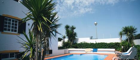 Une piscine dotée d’une terrasse carrelée, entourée de palmiers et bordée d’un bâtiment blanc aux ornements jaunes.