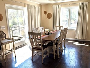 A dining room with a wooden table, four chairs, and two windows with curtains. - New 5BR Luxury Home Near Ferry – Huge Deck, Fire Pit, Sleeps 12 (Tisbury)