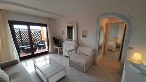A spacious living area with a white sofa, a glass coffee table, and a wicker chair. There is a TV, a small table, and a cabinet in the room. - ELEGANTE BILO D2 CASA SANDRA (Golfo di Marinella)