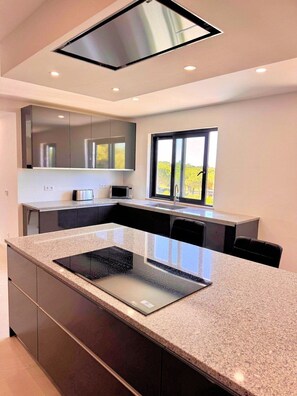 A modern kitchen with a central island, built-in appliances, and a large window providing natural light. - Casa Vilamoura Luxe | Luxury 9BR retreat | Walk to Marina | Vilamoura (Quarteira)