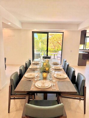 A dining room with a long table set for a meal, surrounded by chairs, and large windows offering a view of trees. - Casa Vilamoura Luxe | Luxury 9BR retreat | Walk to Marina | Vilamoura (Quarteira)