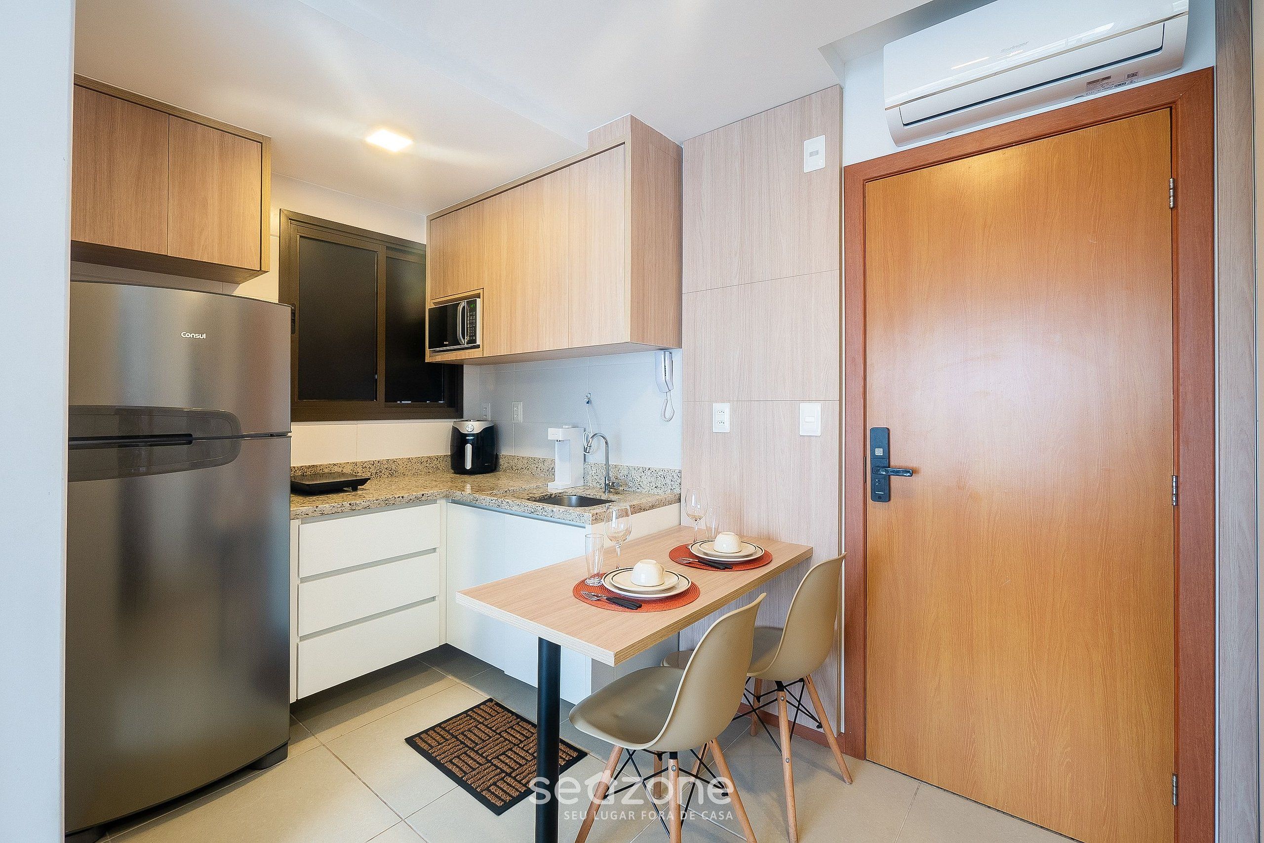 A compact kitchen with a stainless steel refrigerator, a microwave, and a sink. There is a dining table with two chairs and a small rug.