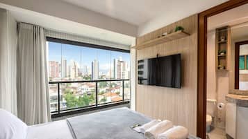 A hotel room with a large window offering a city view, a flat-screen TV mounted on the wall, and a neatly made bed with towels.