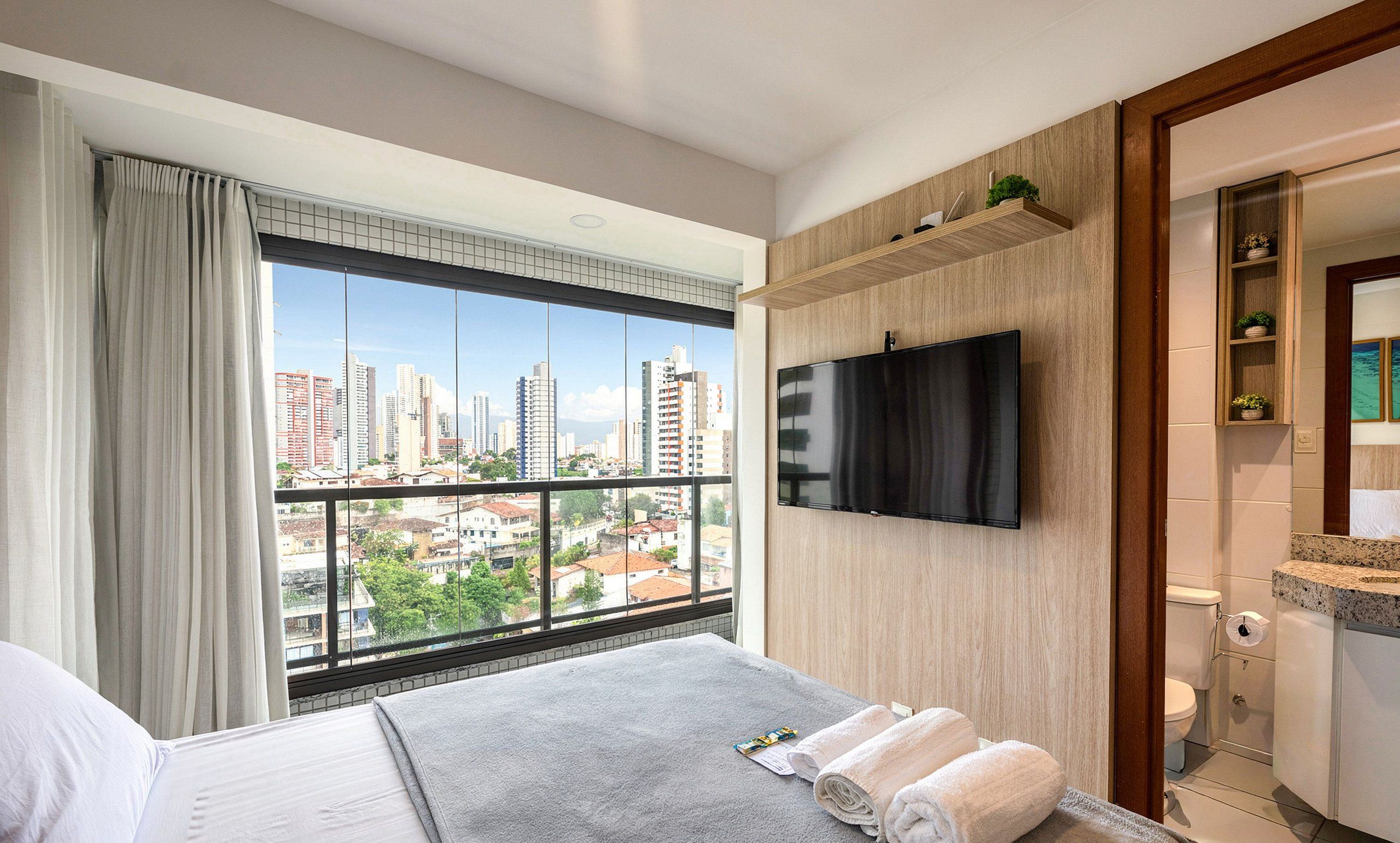 Ein Hotelzimmer mit großem Fenster und Blick auf die Stadt, einem flachbildfernseher an der Wand und einem ordentlich bezogenen Bett mit Handtüchern.