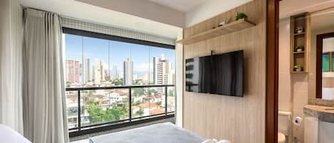 A hotel room with a large window offering a city view, a flat-screen TV mounted on the wall, and a neatly made bed with towels.