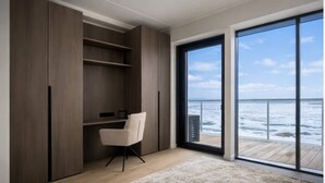 Quarto com uma grande porta de vidro deslizante que dá para uma varanda com vista para o oceano. Há uma cadeira branca e piso de madeira.