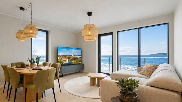 Uma sala de estar moderna com uma grande janela com vista para o mar, uma área de jantar com mesa redonda e um espaço de estar acolhedor com sofá e planta em vaso.