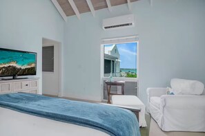 Een slaapkamer met een bed, een televisie, een stoel, een tafeltje en een balkon met uitzicht op zee.