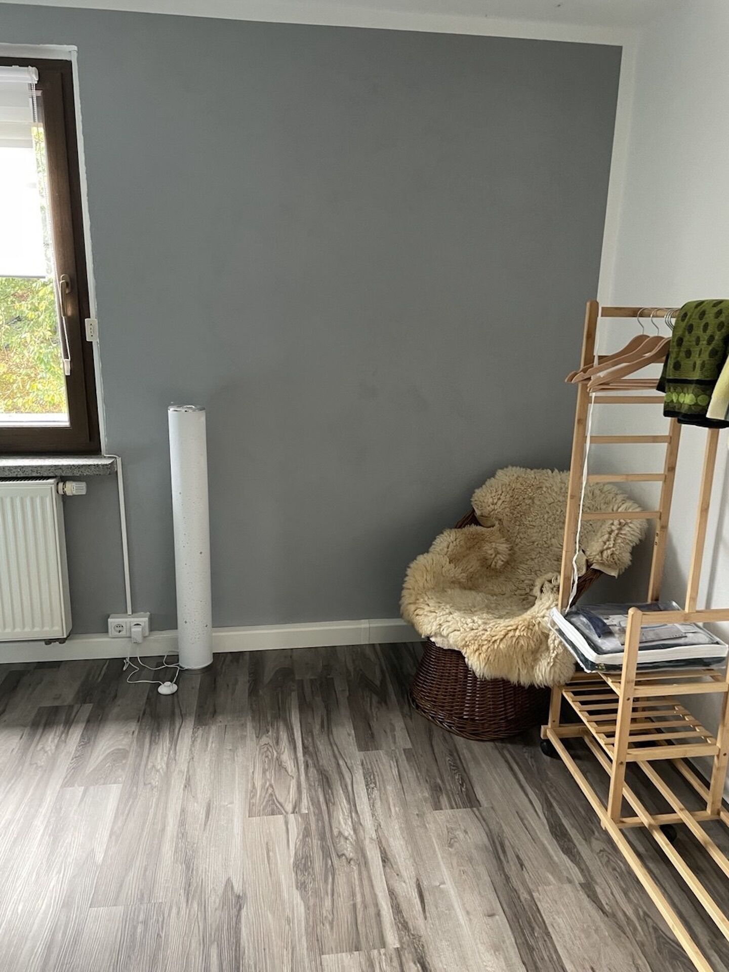 A room with grey walls, a wooden floor, a radiator, a window, a fluffy chair, and a wooden drying rack with clothes.