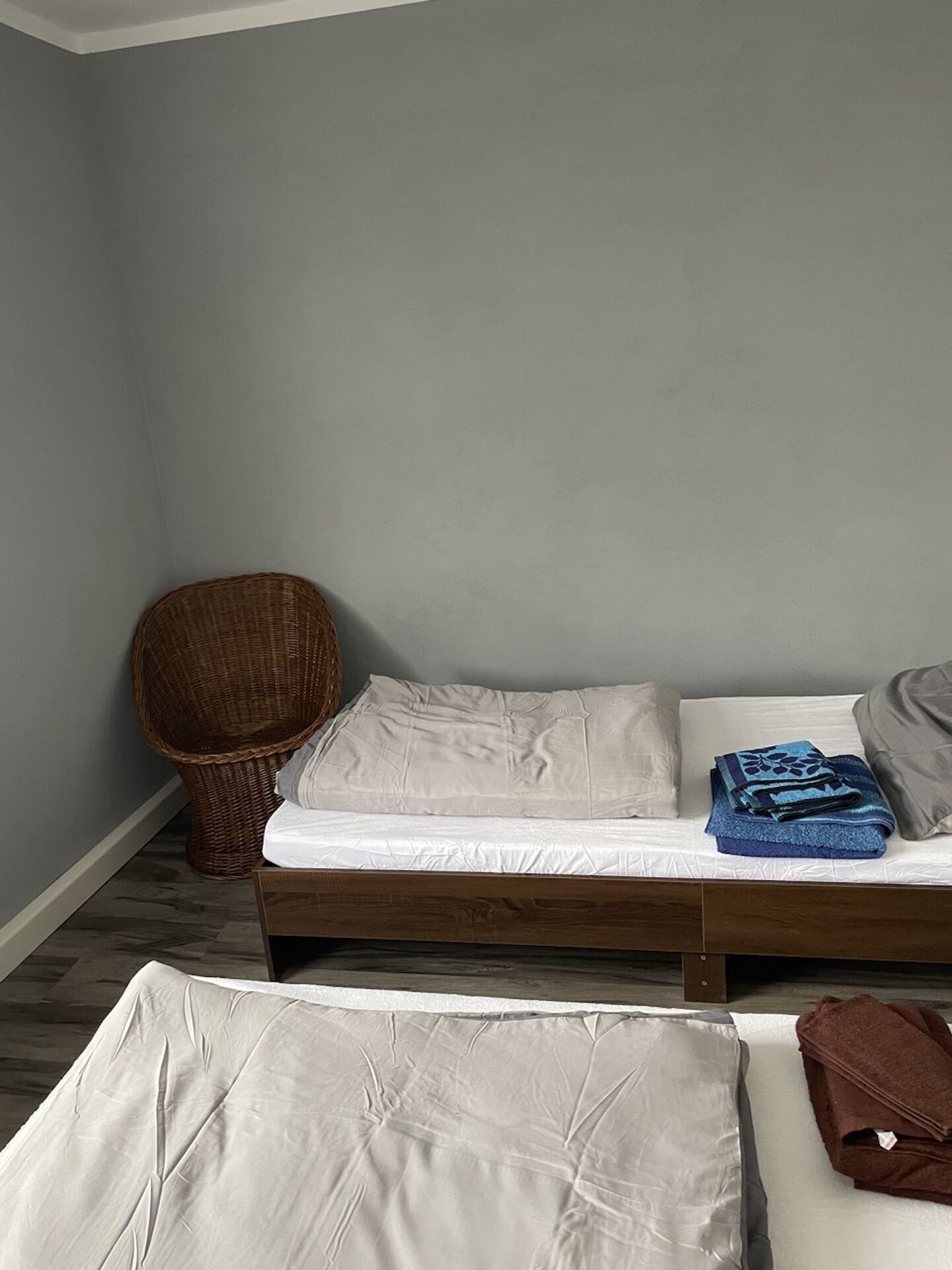 A small, neatly arranged room with a single bed, a wicker chair, and a small table.