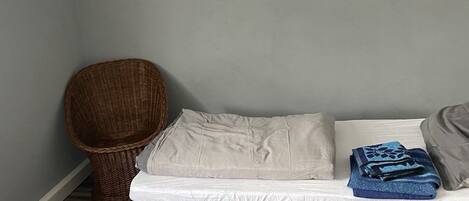 A small, neatly arranged room with a single bed, a wicker chair, and a small table.