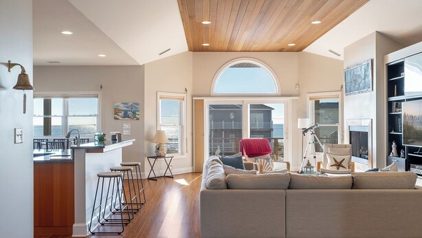 Un soggiorno spazioso con un ampio divano angolare grigio, un soffitto in legno e vista sull'oceano attraverso una grande finestra.