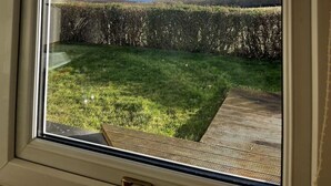 On peut voir, à travers la fenêtre, un espace verdoyant avec une terrasse en bois et une haie.