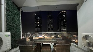 A balcony with wicker furniture, an air conditioning unit, and a view of city buildings at night. - Said's Suites Elysium tower opposite to centaurus mall Islamabad (Islamabad)