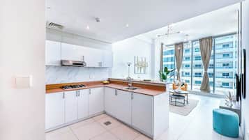 Cozinha moderna com armários brancos, bancada de mármore e fogão a gás. Há uma área de refeições com mesa e cadeiras, e uma grande janela com vista para um edifício.