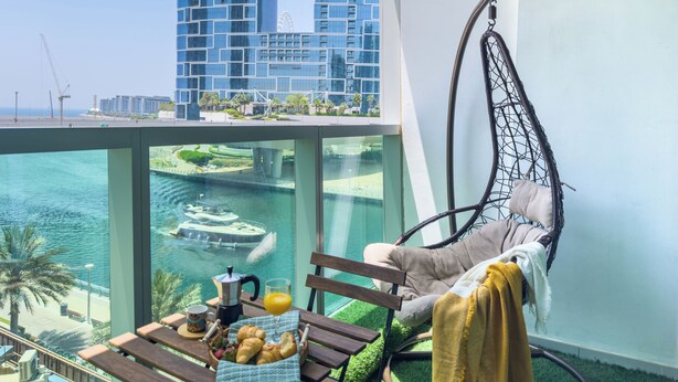A balcony with a hanging chair, a table with breakfast items, and a view of a marina and modern buildings. - Marina View Stay with Rooftop Pool (Dubai)
