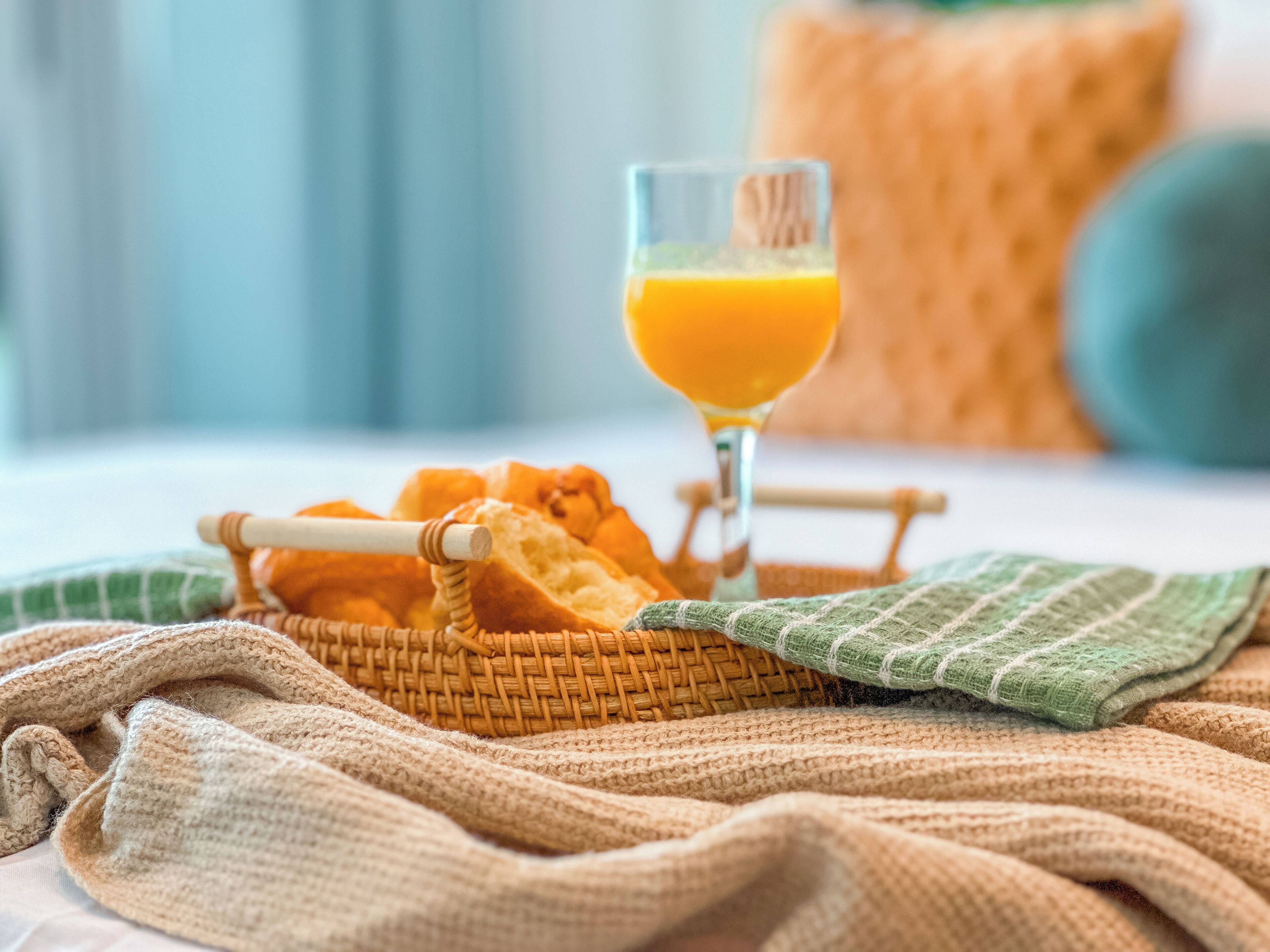 A wicker basket with bread and a glass of orange juice on a bed with a green and beige blanket.