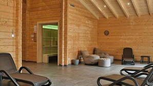 Interior de una cabaña de troncos con techo alto, paredes de madera, una zona de estar con sillas y cojines, y una puerta visible que conduce a otra habitación.