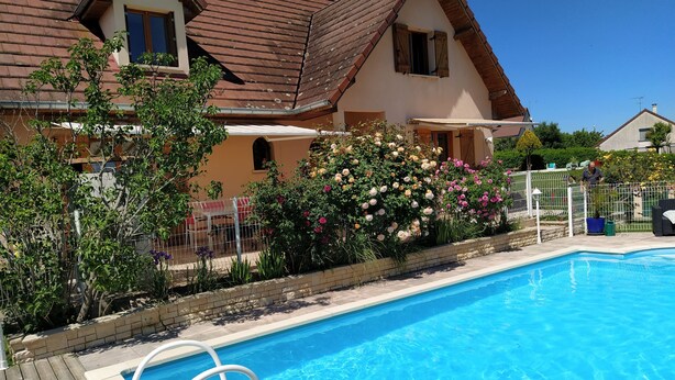 A swimming pool with a wooden deck, surrounded by a house with a sloped roof and flowering bushes. - Summer rental Villa & pool Lons le Saunier (Courlans)