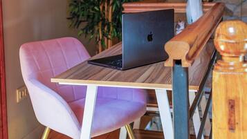 Une table en bois avec un ordinateur portable, une chaise rose et une plante, dans une pièce rustique aux murs de pierre et au plafond en bois.