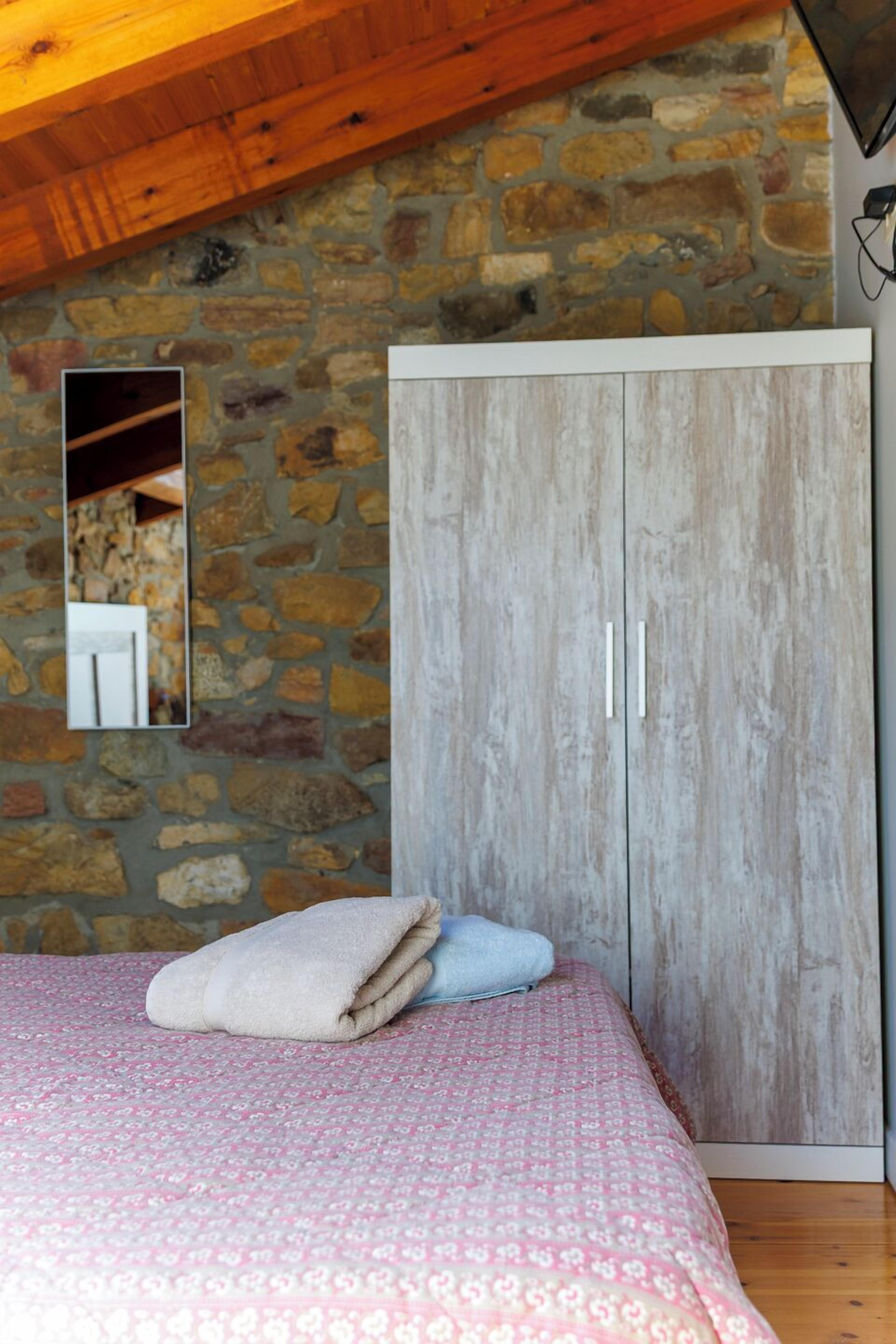 A bedroom with a stone wall, wooden ceiling, a wardrobe, and a bed with a patterned bedspread.