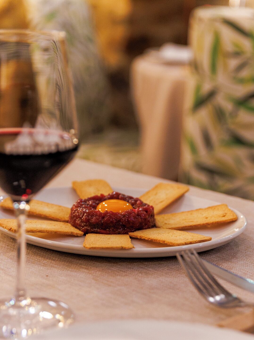 Un plato con un guiso de carne en el centro, rodeado de tostadas, una copa de vino tinto y un tenedor sobre un mantel.