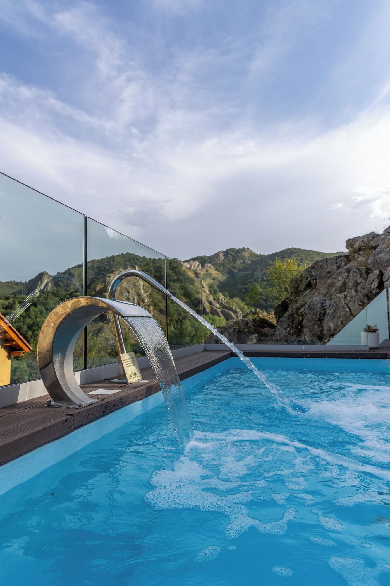 A modern outdoor pool with a waterfall feature, surrounded by a glass railing and overlooking a mountainous landscape.