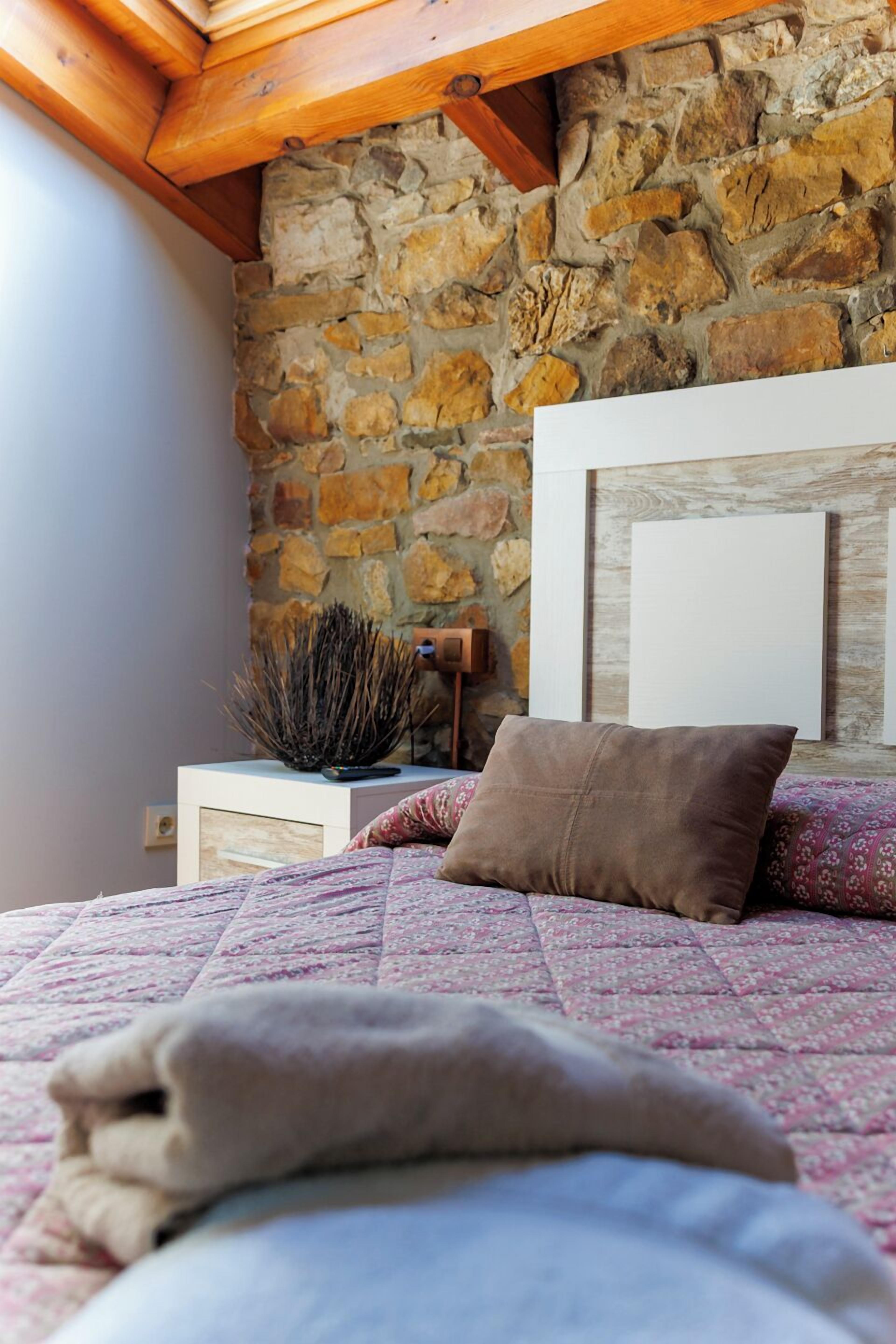 Un dormitorio con pared de piedra, techo de madera, cama con colcha y mesita de noche con lámpara y ramas decorativas.