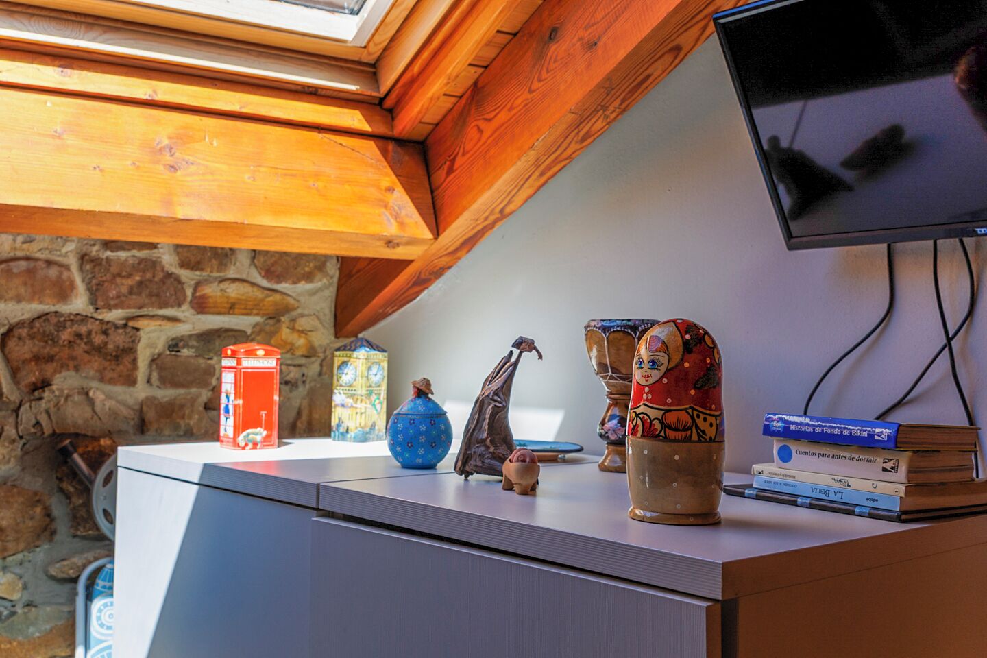 Una habitación con pared de piedra, techo de madera, televisor de pantalla plana y varios objetos decorativos, incluyendo una caja roja, un jarrón azul y una pila de libros.