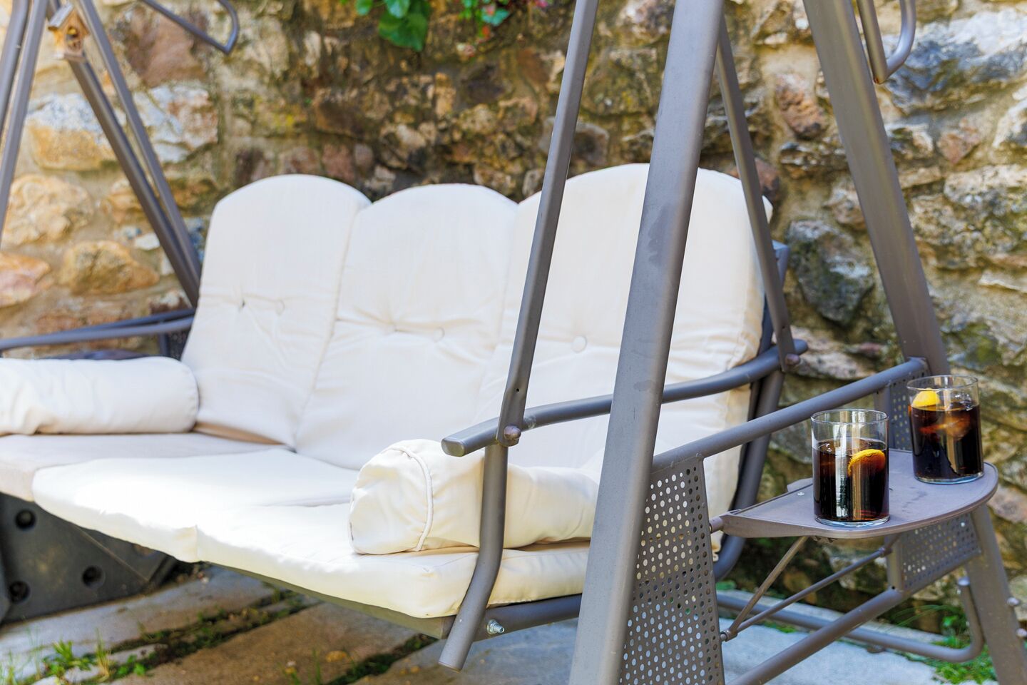 A white cushioned swing chair with a metal frame, two glasses of dark beverage with lemon slices, and a stone wall in the background.