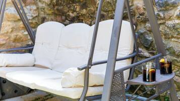 A white cushioned swing chair with a metal frame, two glasses of dark beverage with lemon slices, and a stone wall in the background.