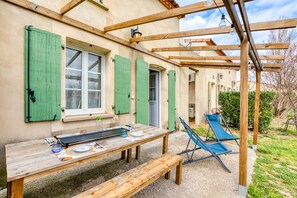 Une table en bois avec un banc, un fauteuil de jardin bleu et une pergola illuminée de guirlandes lumineuses.