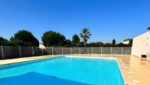 Una piscina rectangular con superficie de agua azul cristalina, rodeada por una cerca blanca y un solárium con baldosas.
