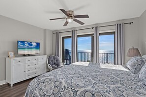 A bedroom with a large bed, a ceiling fan, a TV on a dresser, a chair, and a view of the ocean through large windows. - Unit 403 | Remodeled Fall Gulf-Front Escape—2BR, Balcony & VIP Beach Service (Panama City Beach)