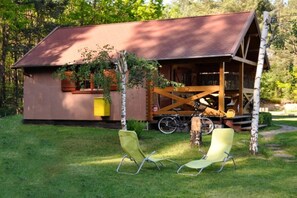 A rustic cabin with a sloped roof, a wooden porch, and a bicycle parked outside. There are two green lawn chairs in front of the cabin. - Summer cottages in Kashubia. (Dąbie)