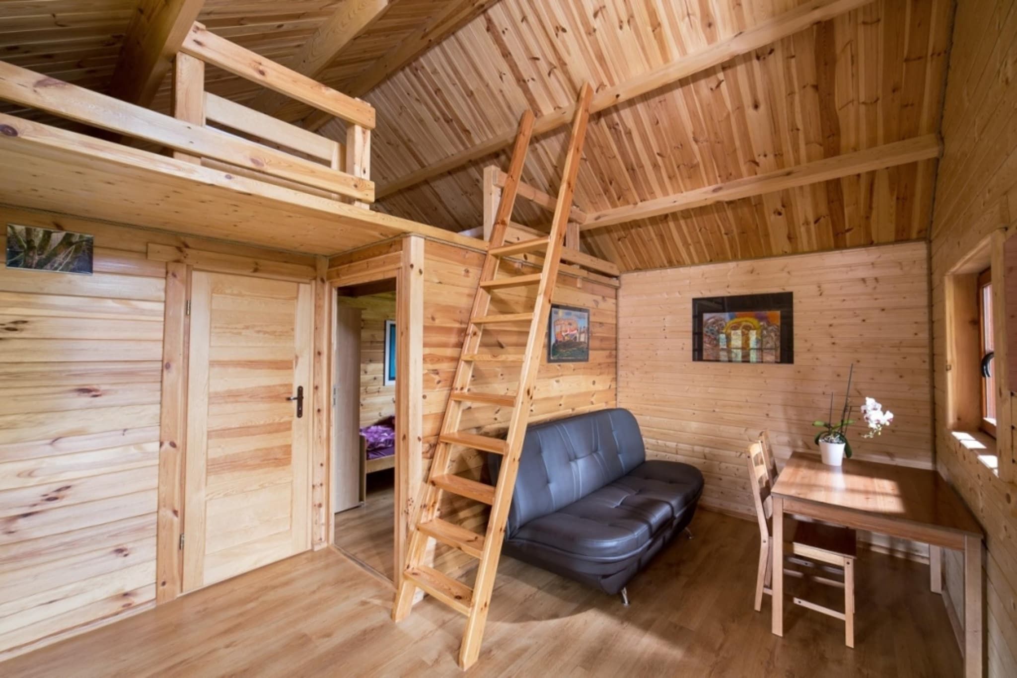 Interior de una cabaña de madera con cama en altillo, sofá, mesa de comedor y escalera.