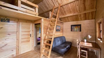 Interior de una cabaña de madera con cama en altillo, sofá, mesa de comedor y escalera.