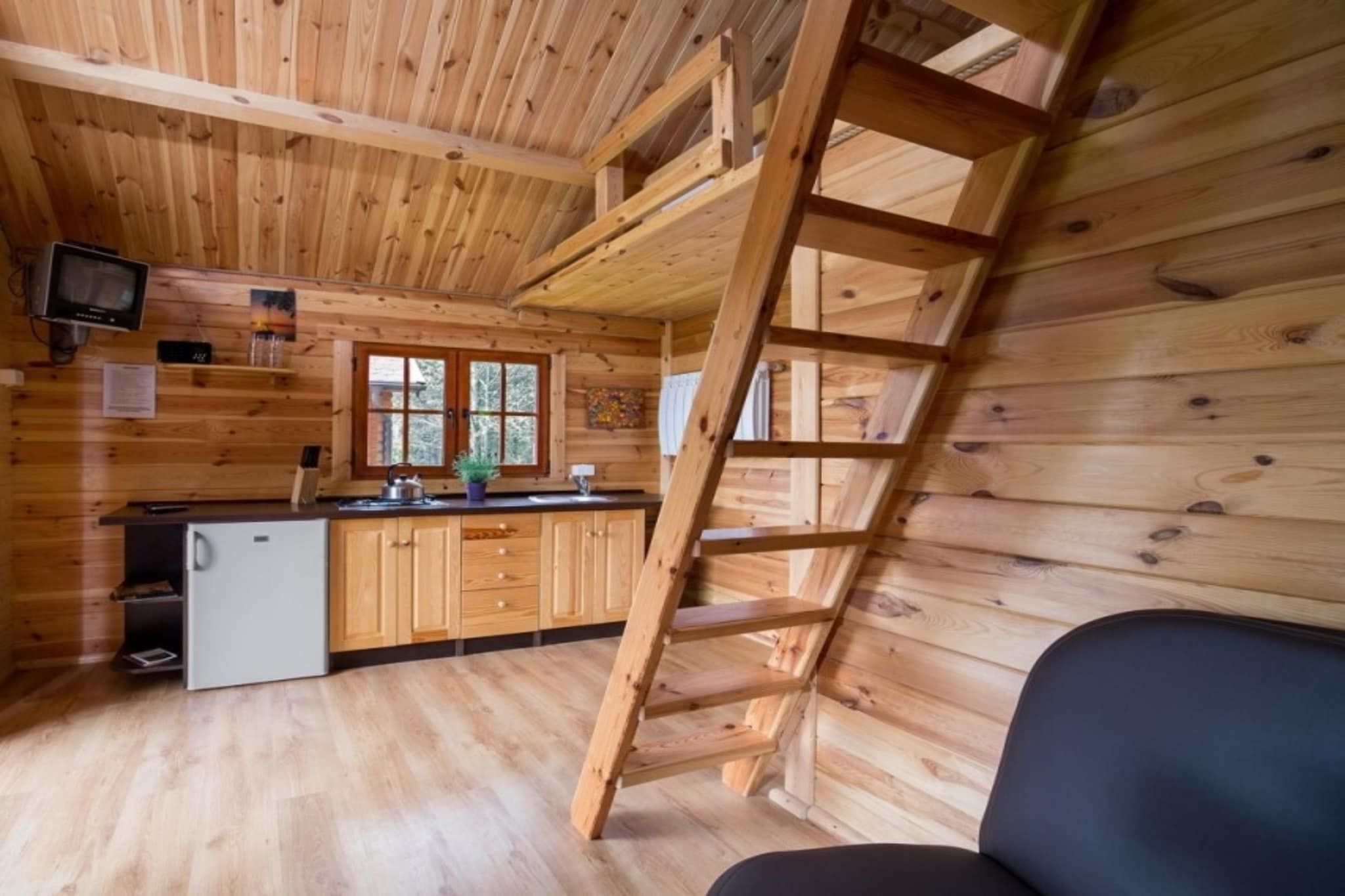 A wooden cabin interior with a kitchen, a ladder, and a small refrigerator.