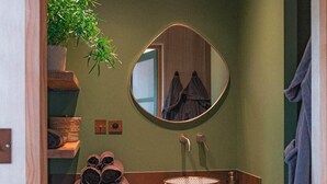 Une salle de bain avec un mur vert, des étagères en bois, un miroir rond, un lavabo avec un récipient tressé et une plante en pot.