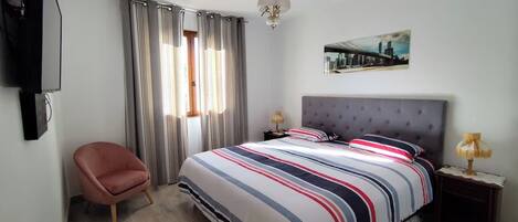 Ein Schlafzimmer mit Bett, Sessel, Fernseher und einem Fenster mit Vorhängen.