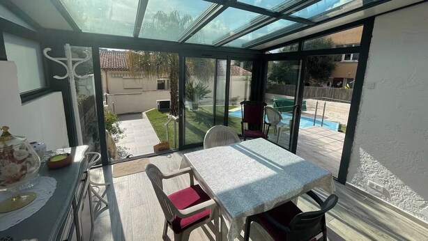 Um pátio ensolarado com mesa e cadeiras, um espaço envidraçado e vista para uma piscina e jardim.