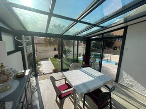 Une terrasse ensoleillée avec une table et des chaises, une véranda et une vue sur une piscine et un jardin.