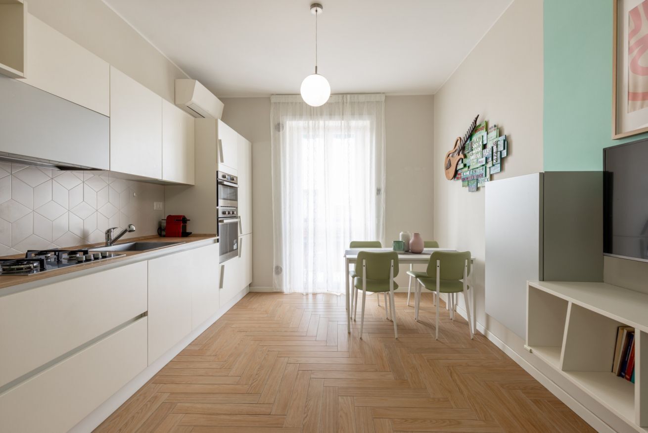 Een moderne keuken met witte kasten, een visgraatvloer, een eethoek met een tafel en stoelen, en een televisie aan de rechterkant.