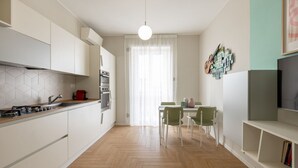 A modern kitchen with white cabinets, a herringbone floor, a dining area with a table and chairs, and a TV on the right. - BDC - Tender 2Bdr apartment @Navigli area (Milan)