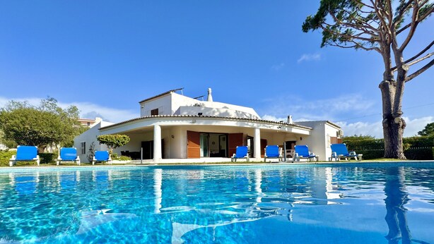 Vista a la piscina de una villa blanca con porche cubierto y sillones reclinables azules.