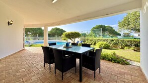Un patio cubierto con una mesa y sillas de vidrio, una piscina al fondo y vistas de la vegetación circundante.