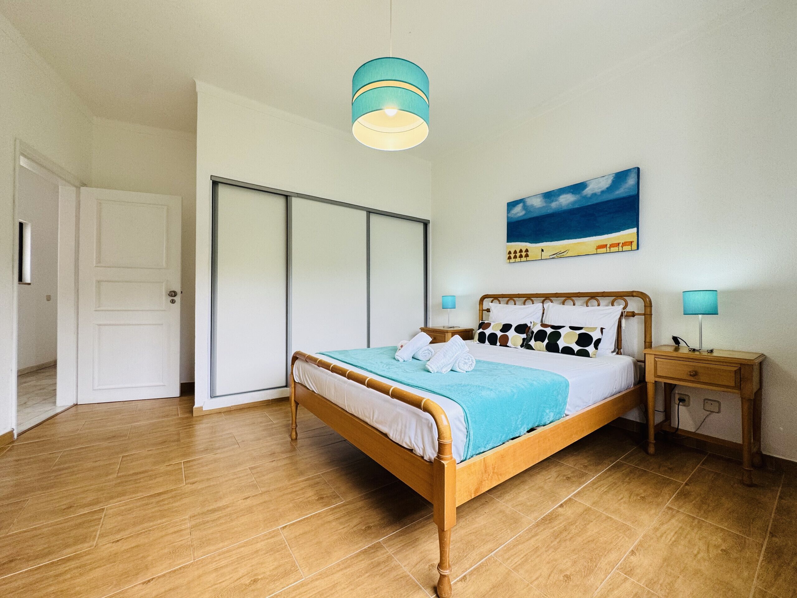A bedroom with a wooden bed, a nightstand, a wall-mounted lamp, a painting of a beach scene, and a sliding door.