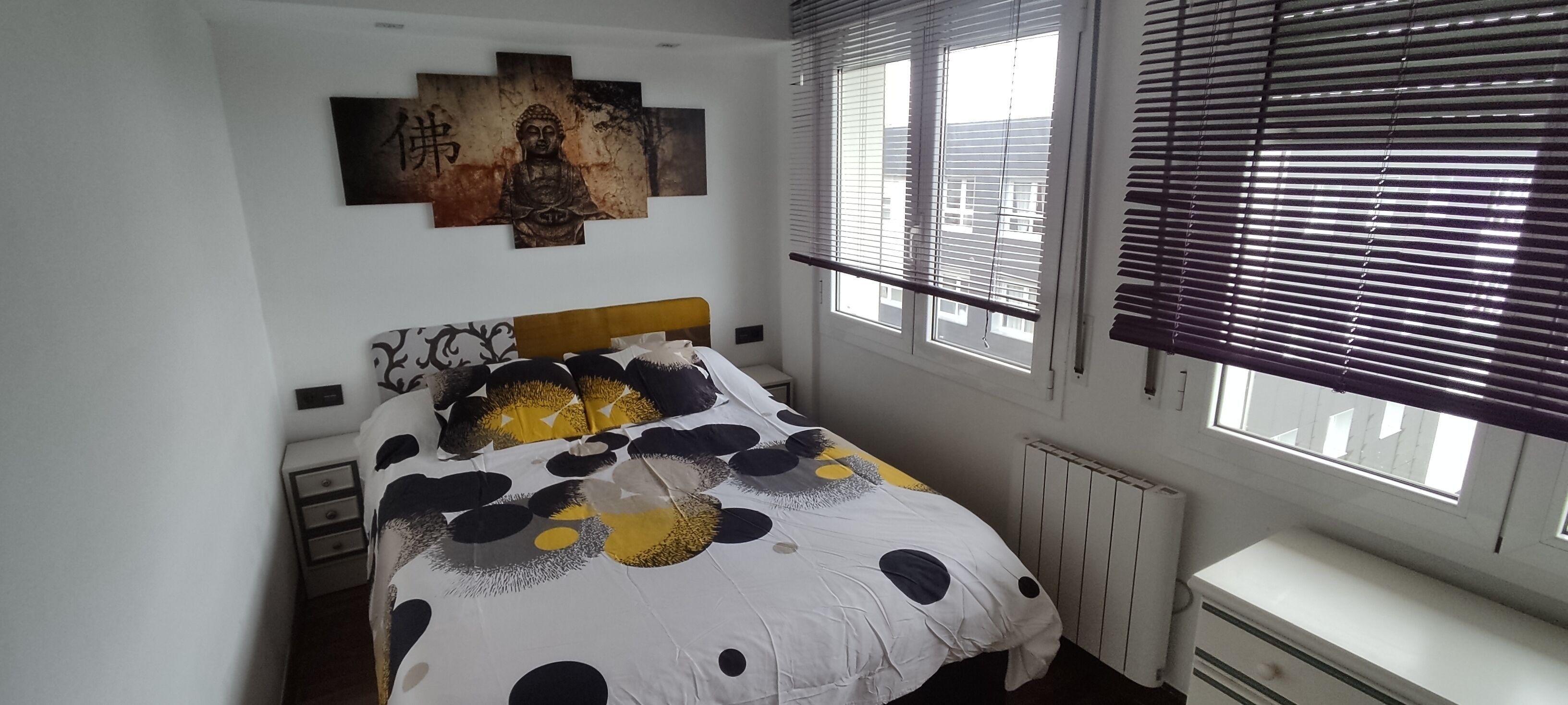 Un dormitorio con una cama, una mesita de noche, un aparador, una ventana con persianas y un cuadro de Buda en la pared.
