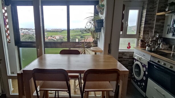 Una cocina con mesa y sillas de madera, lavadora y vistas al campo a través de amplios ventanales.