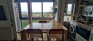 Een keuken met een houten tafel en stoelen, een wasmachine, en uitzicht op het landschap door grote ramen.